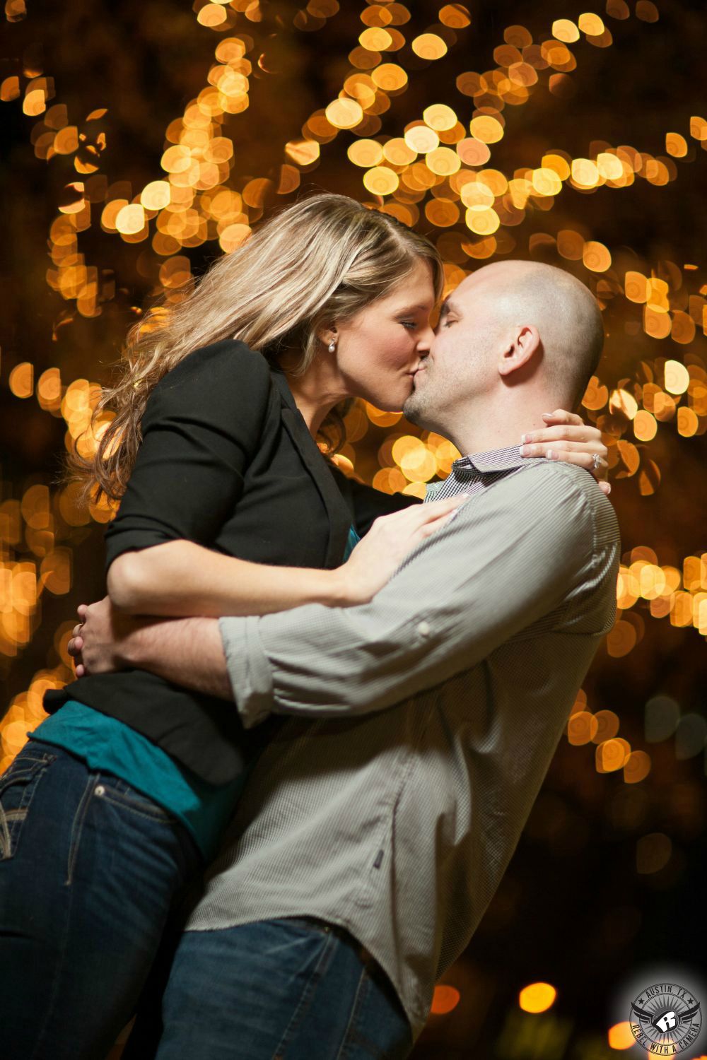 engagement couple with blond haired female with black blazer and blue jeans  and a male groom with grey button up shirt kiss in front of a tree covered with christmas lights causing a bokeh effect on congress avenue in austin	